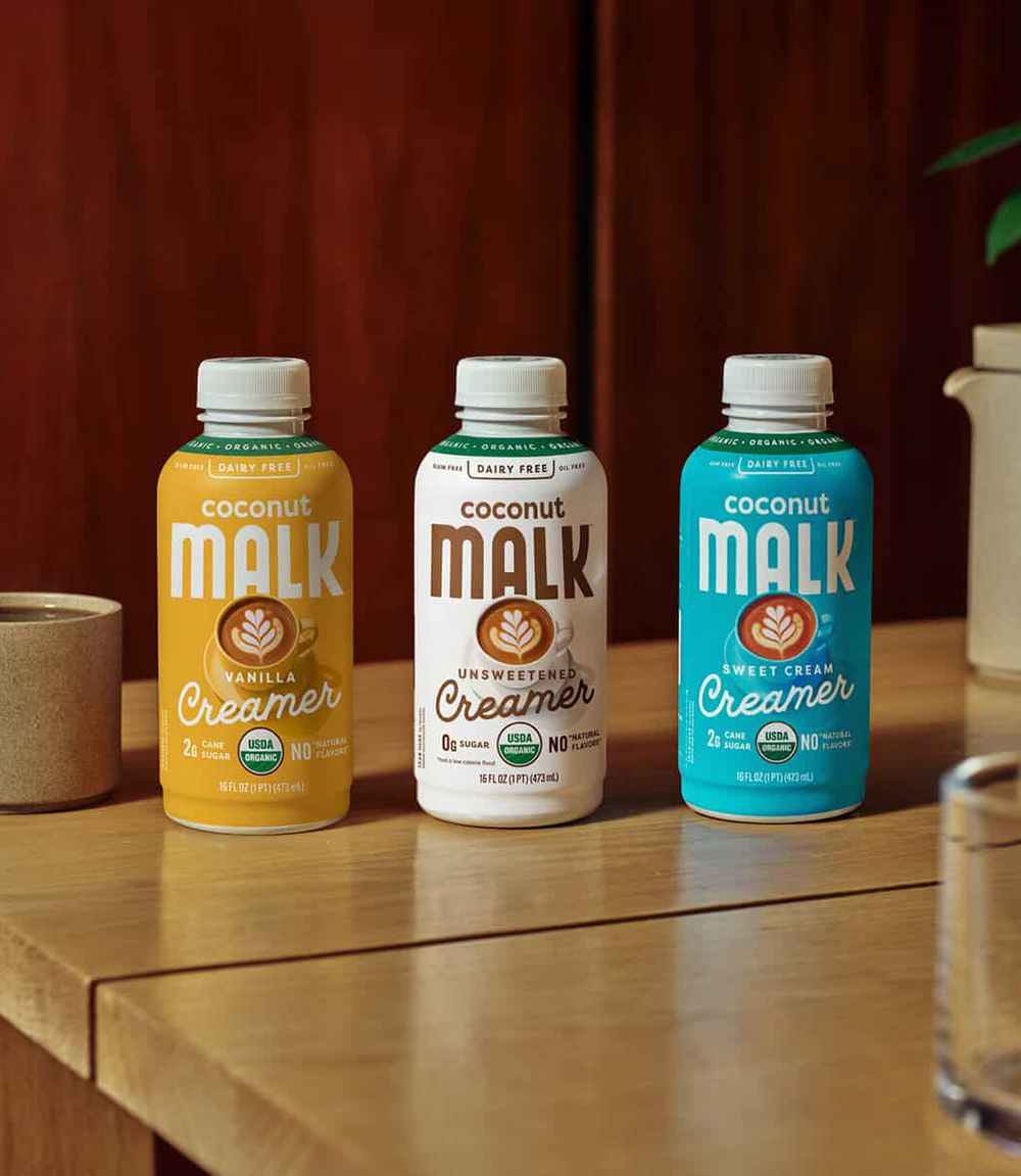 Three bottles of MALK Organic Coconut Creamers in Vanilla, Unsweetened, and Sweet Cream flavors displayed on a wooden table.