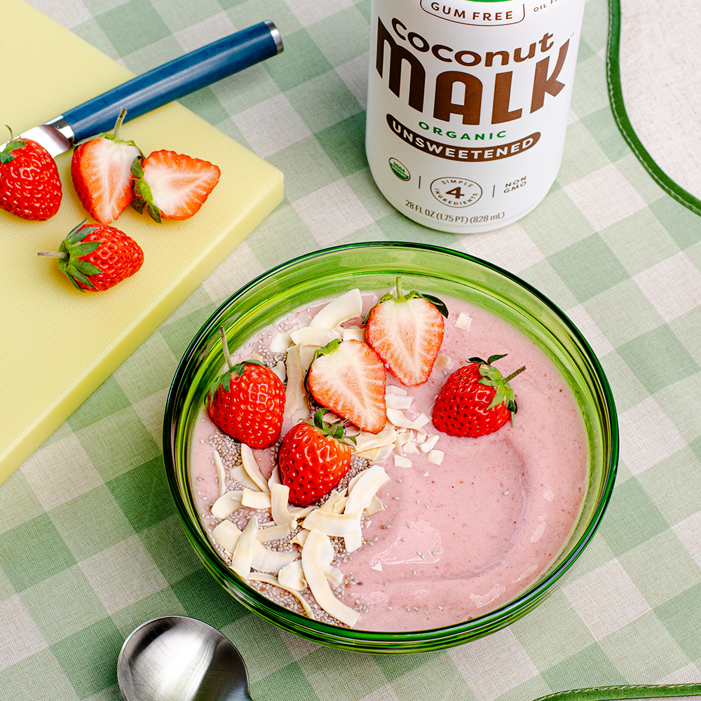Strawberry banana smoothie bowl with fresh strawberries, coconut flakes, and chia seeds made with MALK Organic Coconut Milk on a green checkered table setting