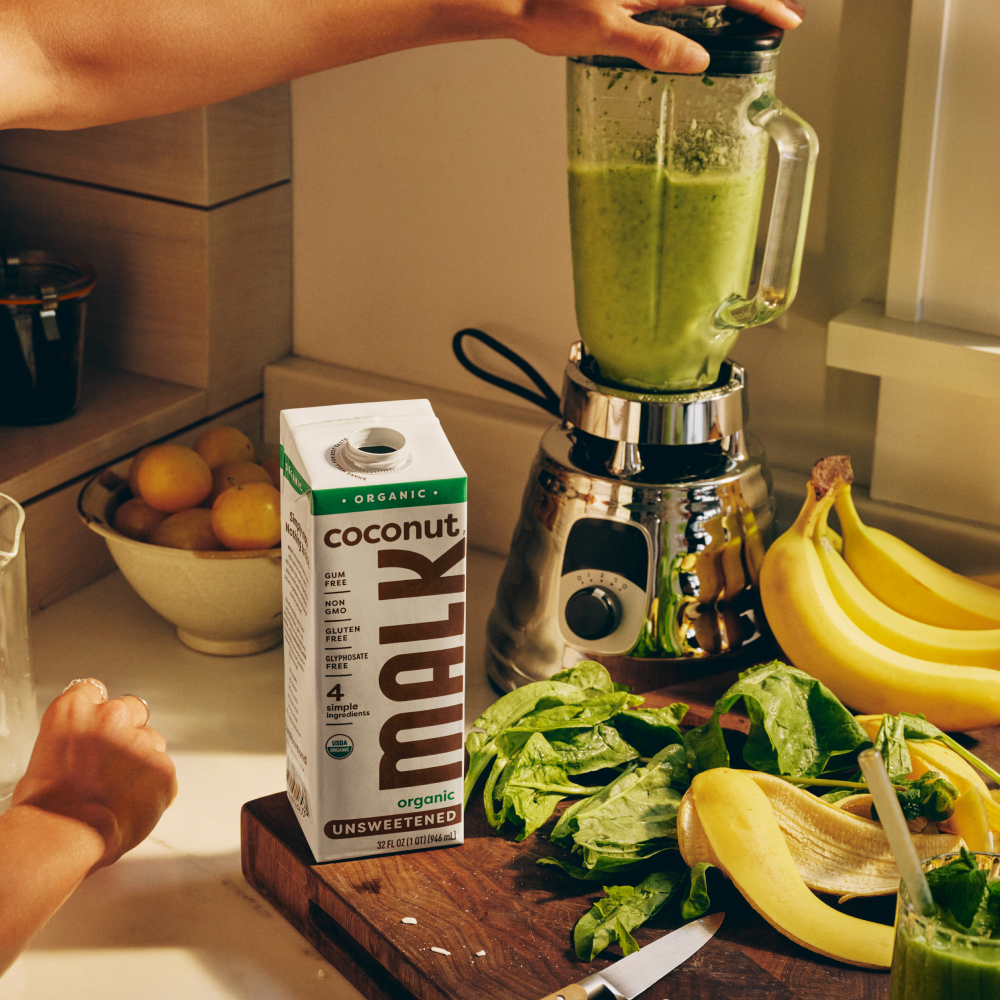 A carton of Shelf Stable MALK Unsweetened Coconut Milk on a kitchen counter beside a blender filled with green smoothie, bananas, spinach, and a hand reaching in the scene.