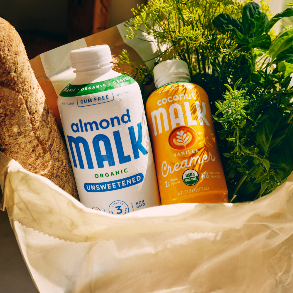 Bottles of MALK Unsweetened Almond Milk and MALK Vanilla Coconut Creamer in a paper bag, with a loaf of bread and green herbs around them.