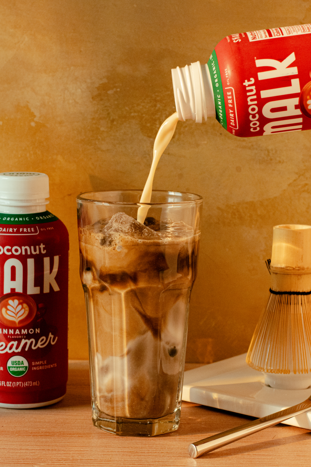 A bottle of MALK Coconut Creamer is shown being poured into a tall glass of iced Hojicha, with another bottle standing on the left and a bamboo whisk on a tray to the right, set against a warm textured background.