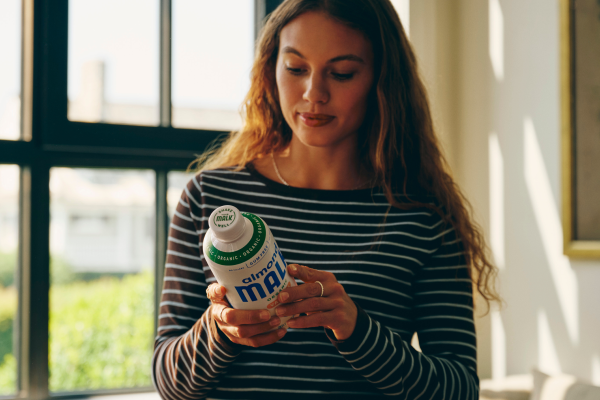 A woman in a striped long-sleeve shirt holds a white bottle of MALK Vanilla Almond Milk near a window, with warm sunlight casting shadows on the wall.