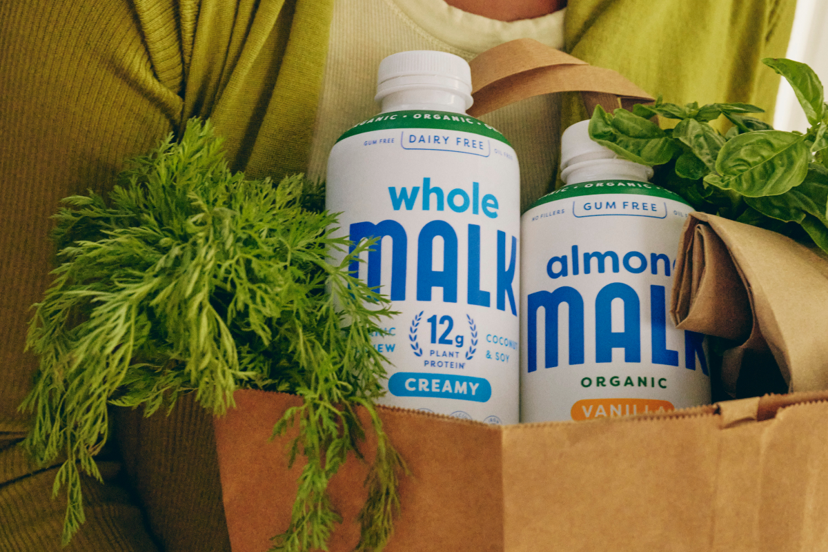 Bottles of Whole MALK and Vanilla Almond MALK in a paper shopping bag with fresh greens shown in a lifestyle setting with a person holding the bags.