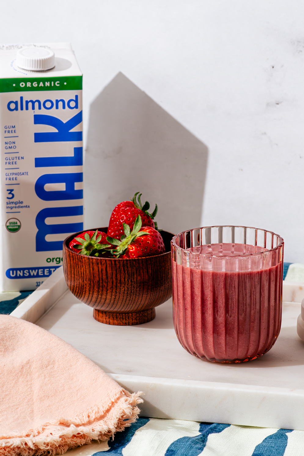 Vanilla berry lime smoothie in a glass next to a bowl of strawberries and a shelf stable carton of Organic Unsweetened Almond MALK