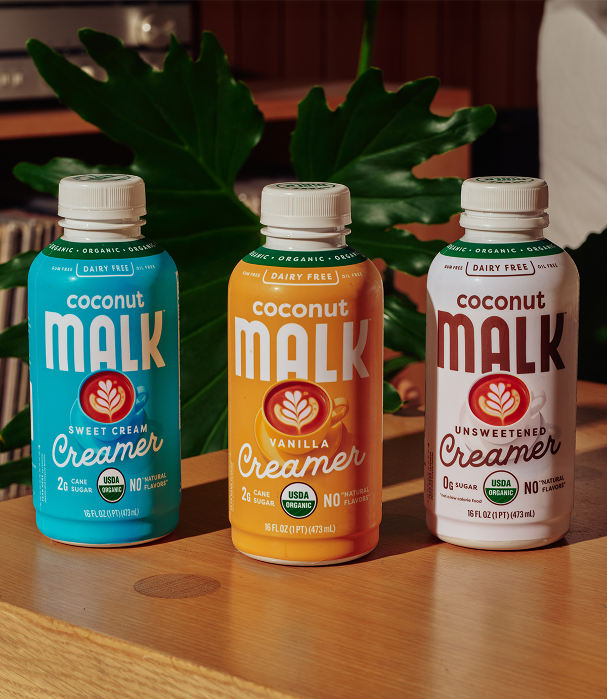 Three bottles of MALK organic coconut-based milk creamers—Sweet Cream, Vanilla, and Unsweetened—displayed on a wooden table, dairy-free plant-based coffee creamers made with simple ingredients.