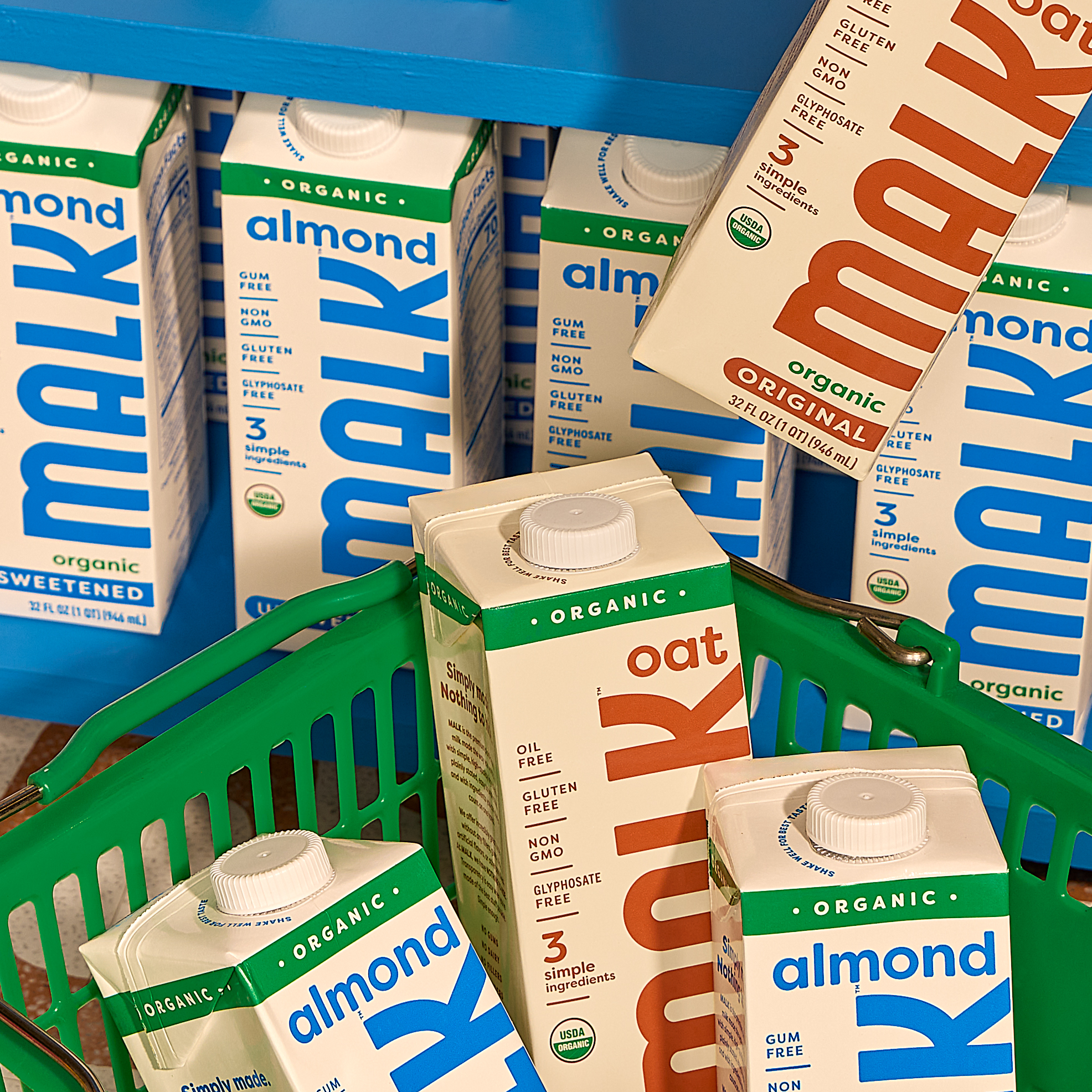 MALK Organic Unsweetened Almond and Original Oat shelf-stable milk cartons displayed in a green shopping basket with more MALK cartons stocked on a blue grocery shelf behind them.