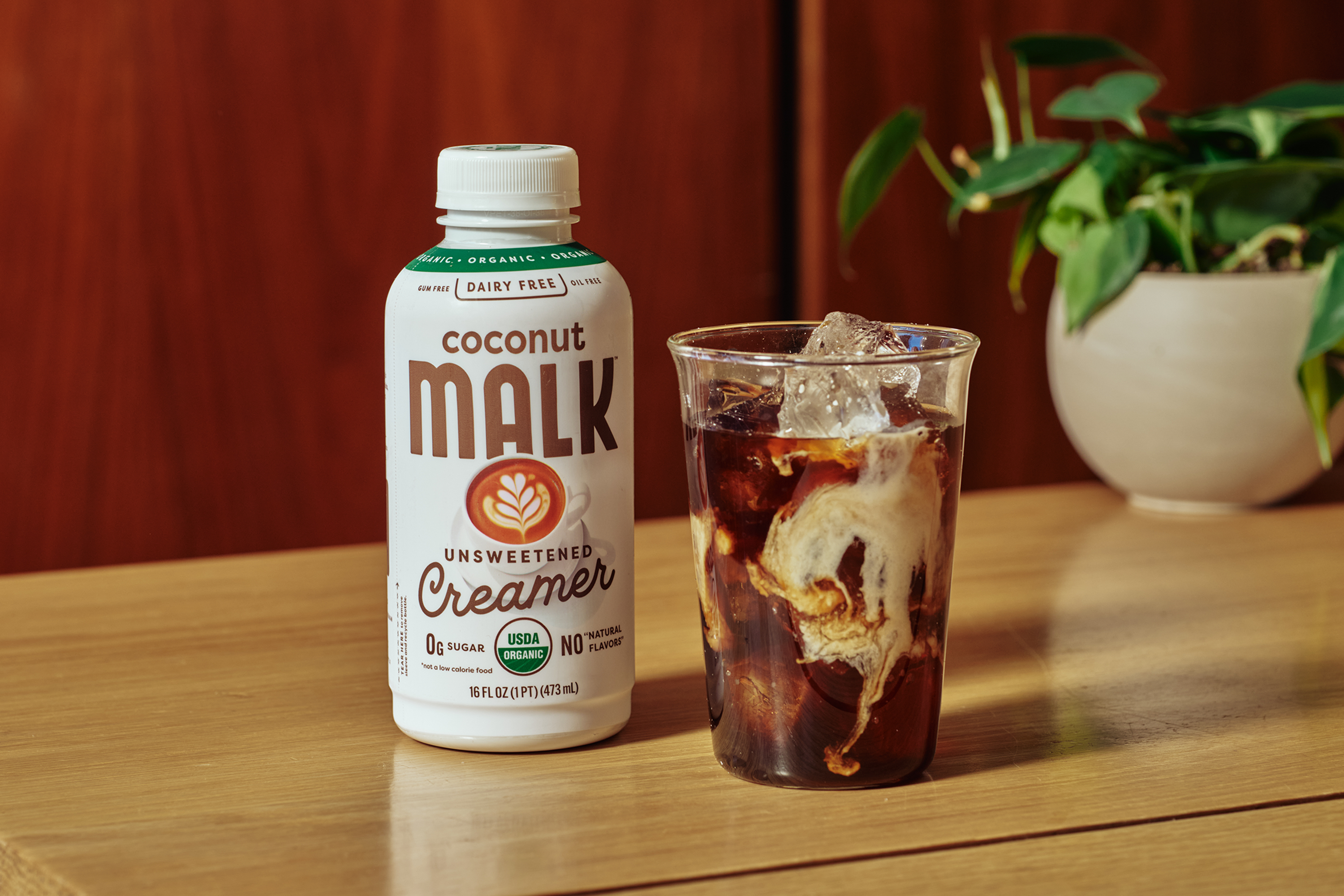 MALK Unsweetened Coconut Milk Coffee Creamer bottle next to a glass of iced coffee with coconut milk swirling, on a wooden table.