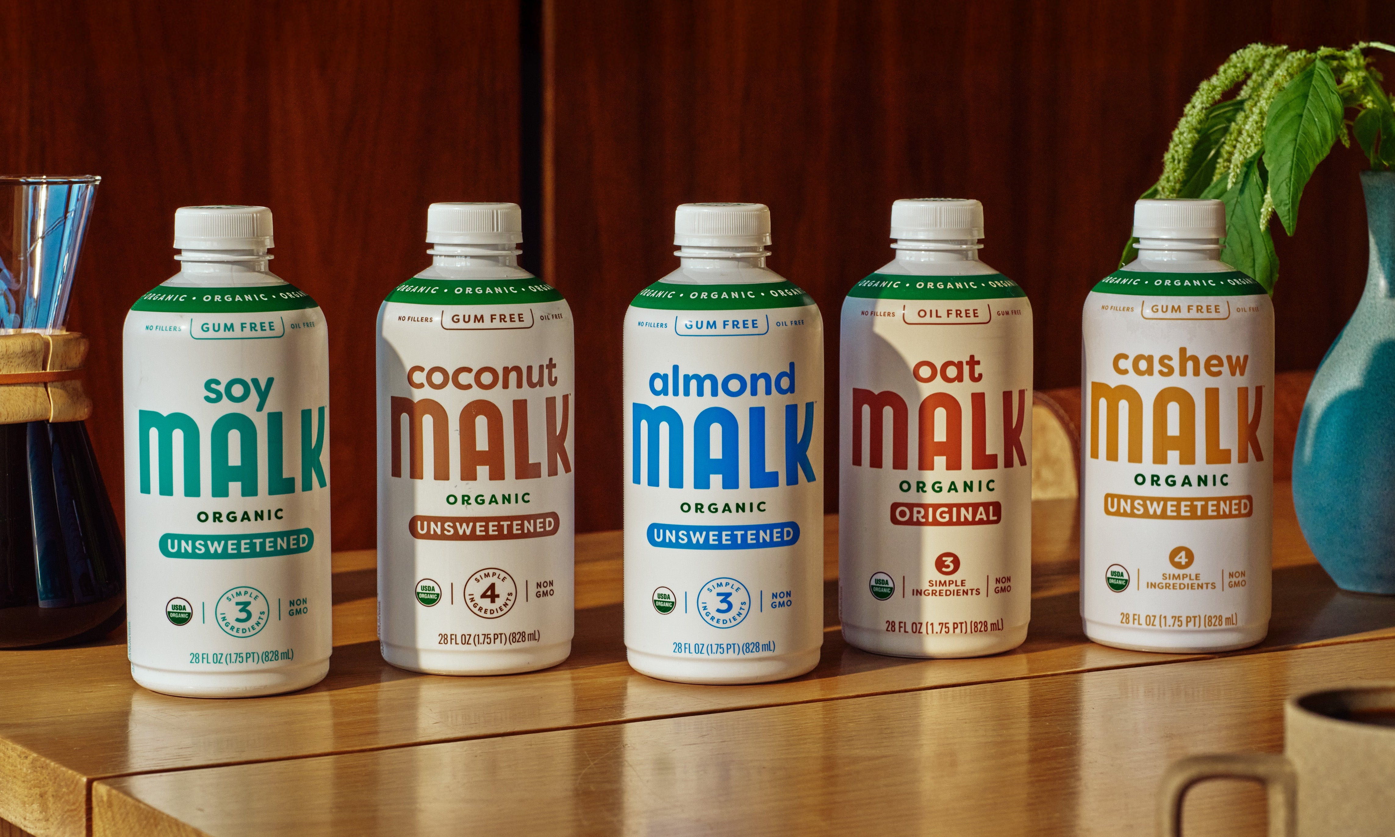 Five bottles of different types of MALK milk alternatives on a wooden surface with a plant and mug in the background.