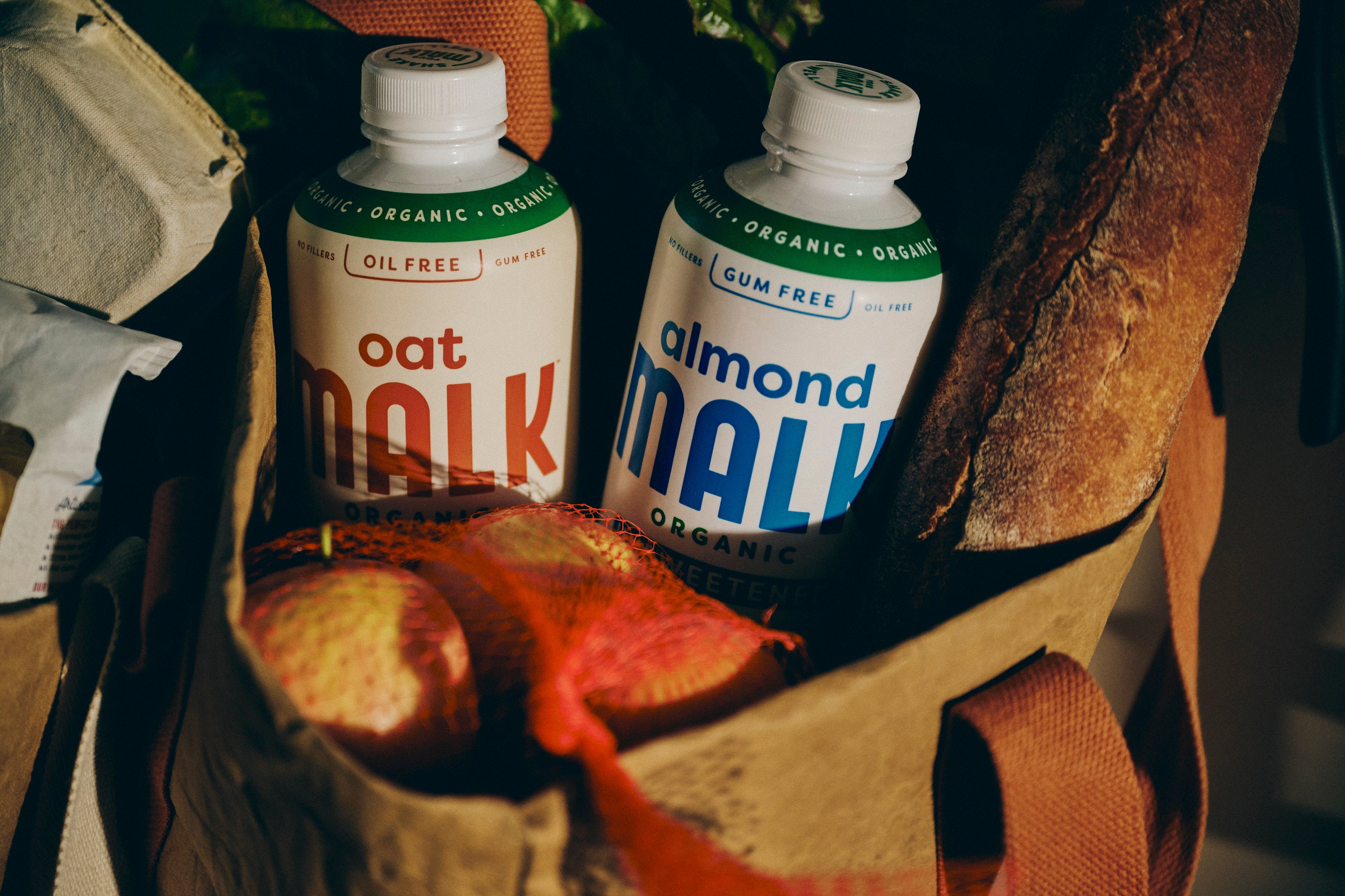 A bottle of oat MALK and bottle of Almond MALK in reusable grocery bag next to a baguette and eggs