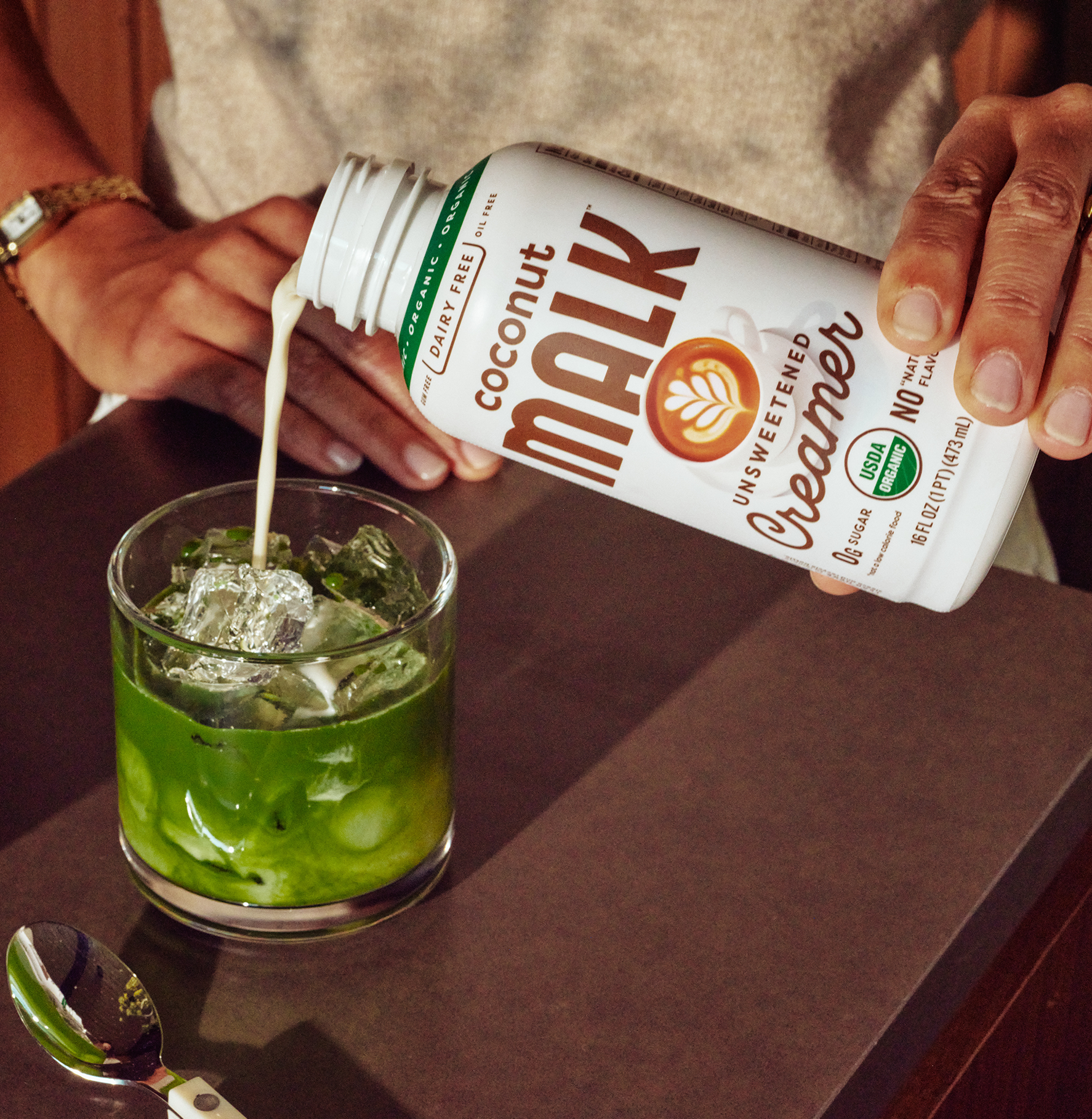 Pouring MALK Unsweetened Coconut Creamer into an iced green matcha drink, showcasing a dairy-free, organic plant-based creamer.