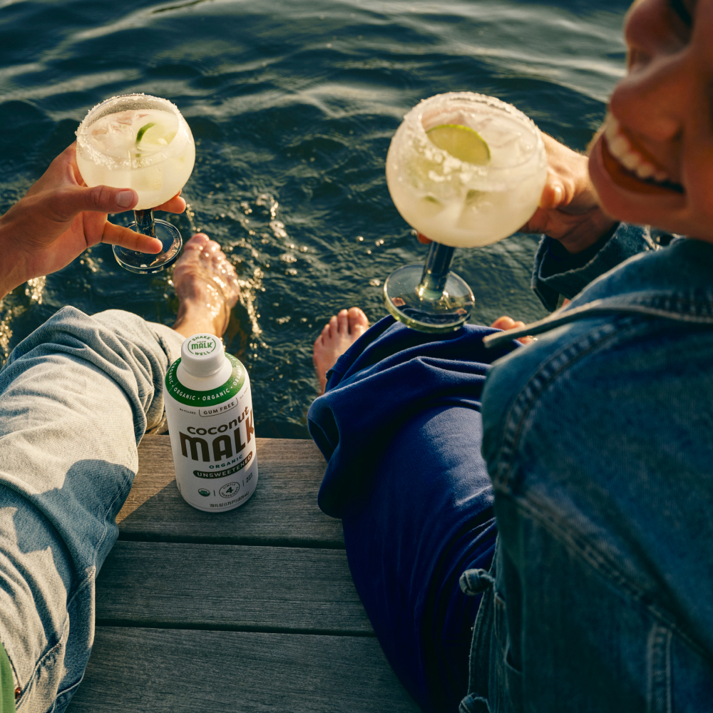Two people sitting on a wooden dock by the water, holding margarita glasses with lime wedges, with a bottle of MALK Unsweetened Coconut Milk on the dock between them.