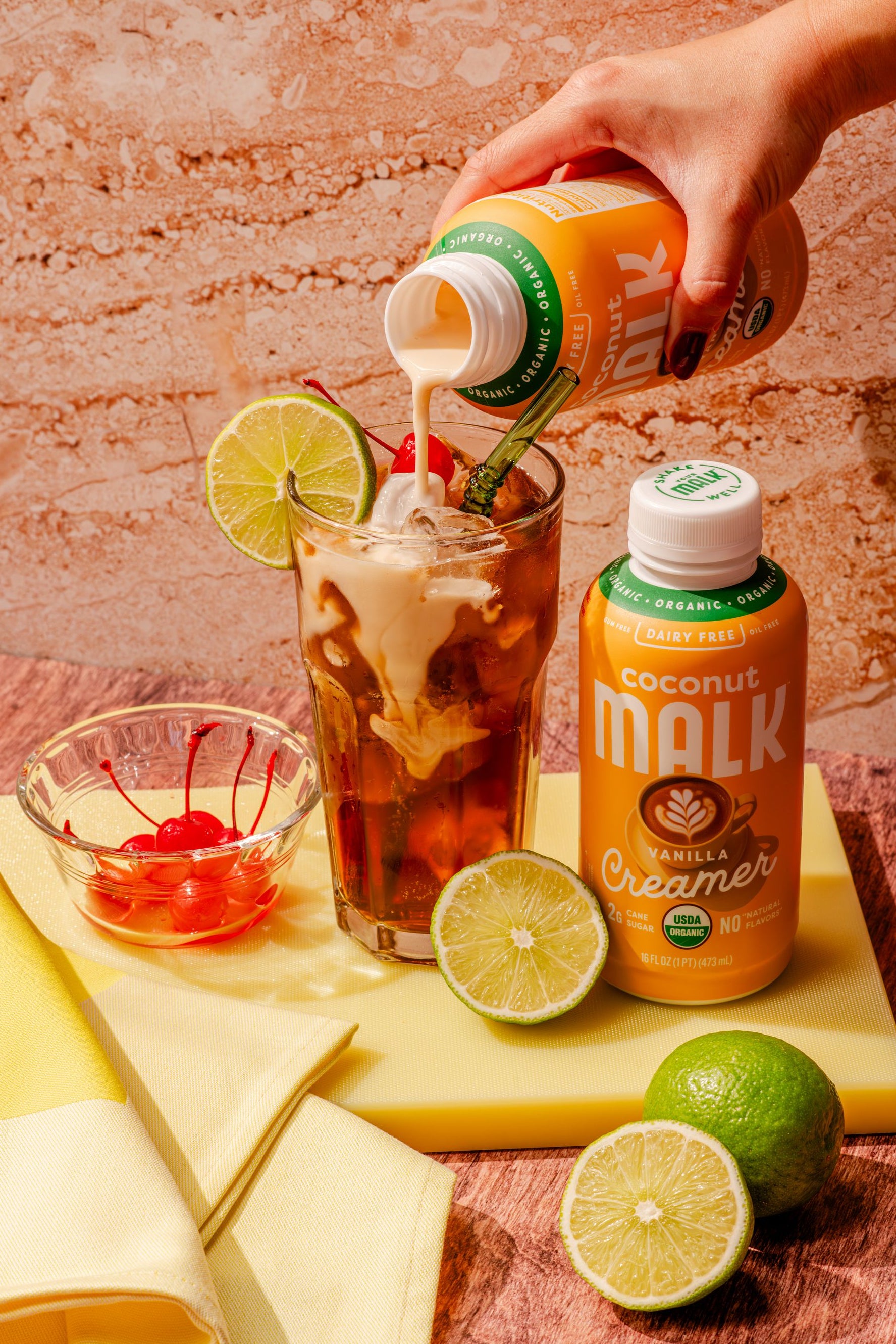 MALK Organic Vanilla Coconut Creamer being poured into a dirty soda with cola, fresh lime, ice, and maraschino cherries.
