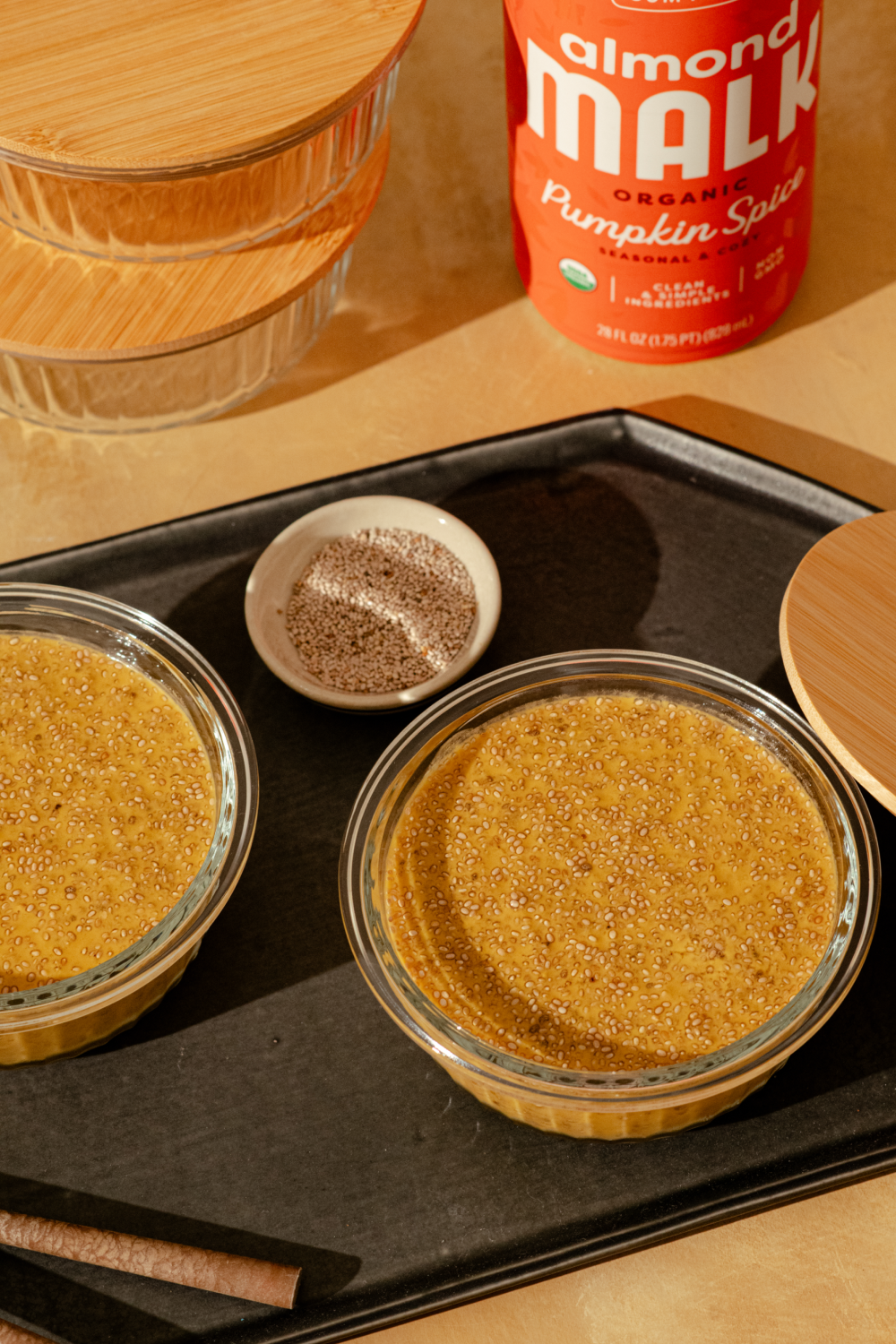 Two glass bowls filled with MALK Pumpkin Spice chia pudding arranged on a black tray, with a small dish of cinnamon and a bottle of Pumpkin Spice MALK in the background.