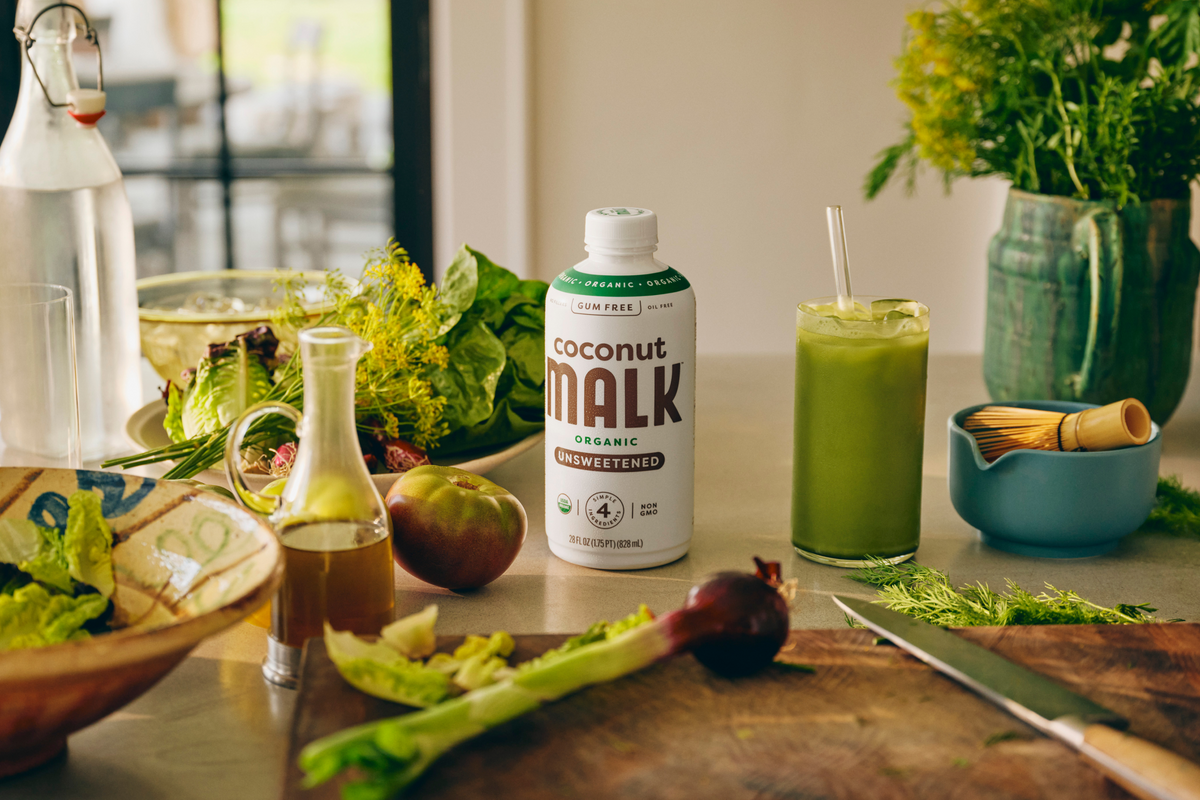 A bottle of MALK Unsweetened Coconut Milk stands on a kitchen counter, surrounded by fresh ingredients and a prepared green beverage with a straw. A salad bowl, cutting board, knife, vase of herbs, and other kitchen items are visible in a bright, real environment.